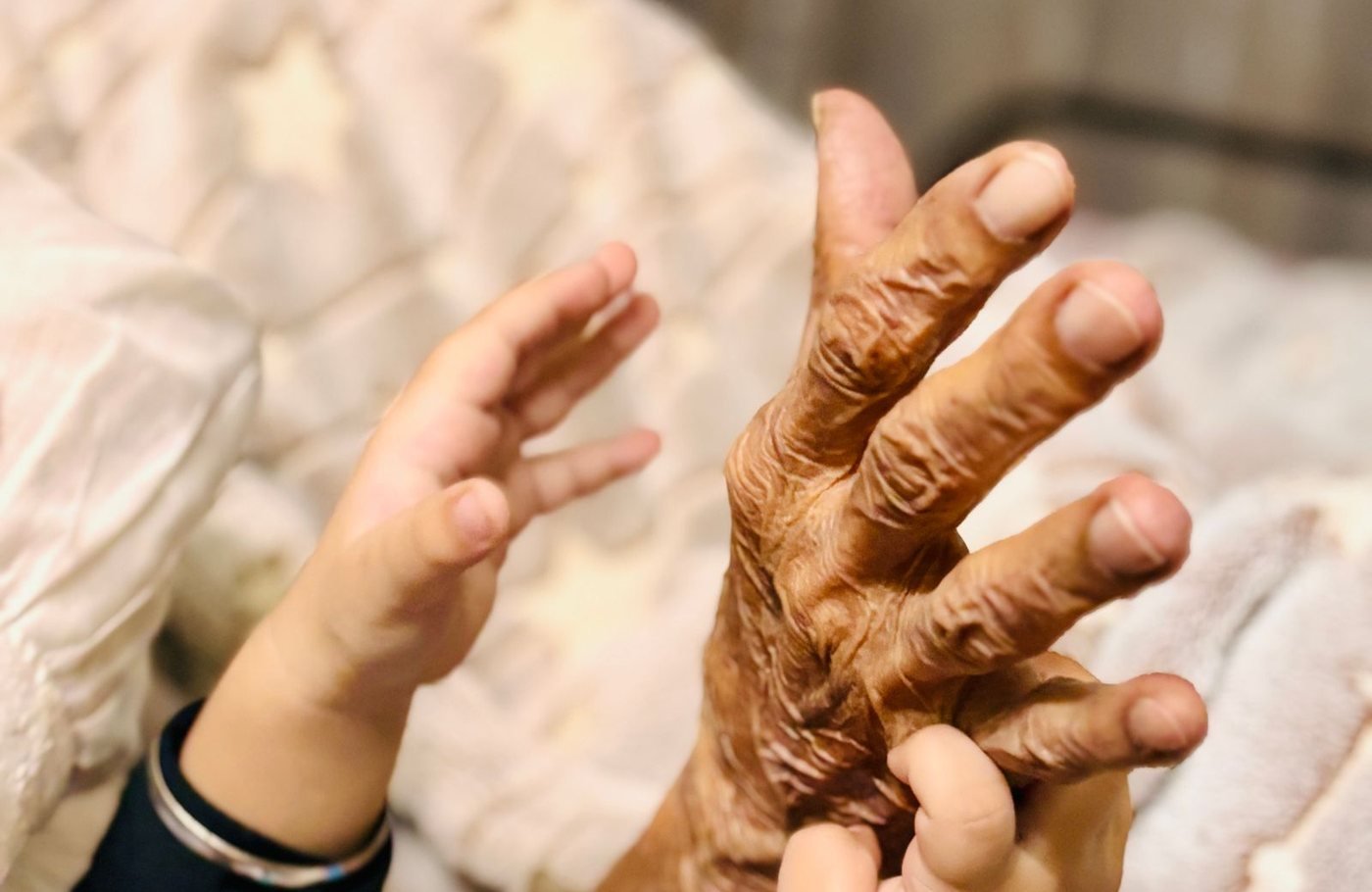 An older adult's hand held gently in a child's hands, resting on a blanket at home.