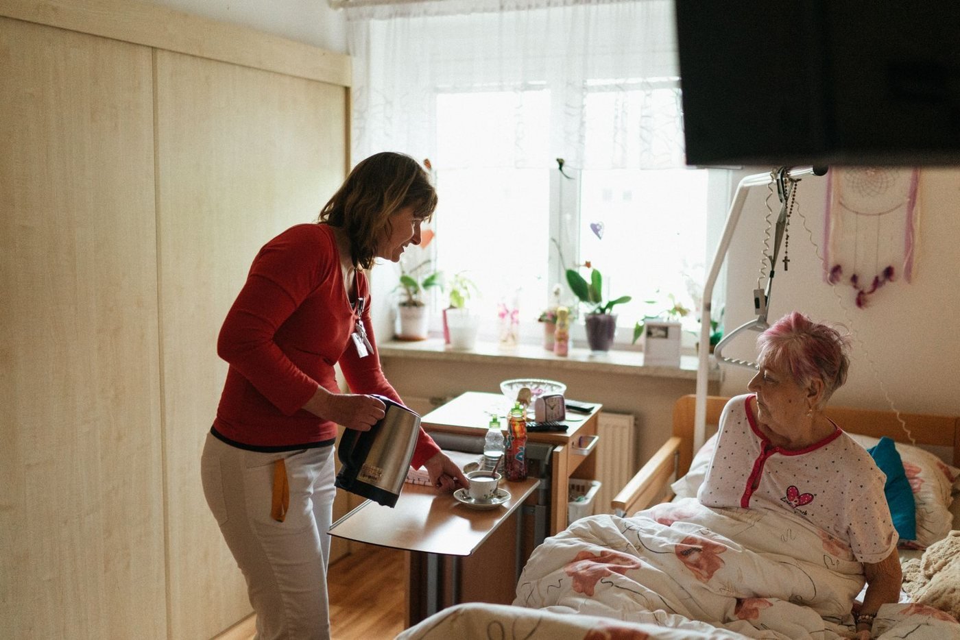 A home caregiver bringing tea to an older adult resting in a sunlit room at home.